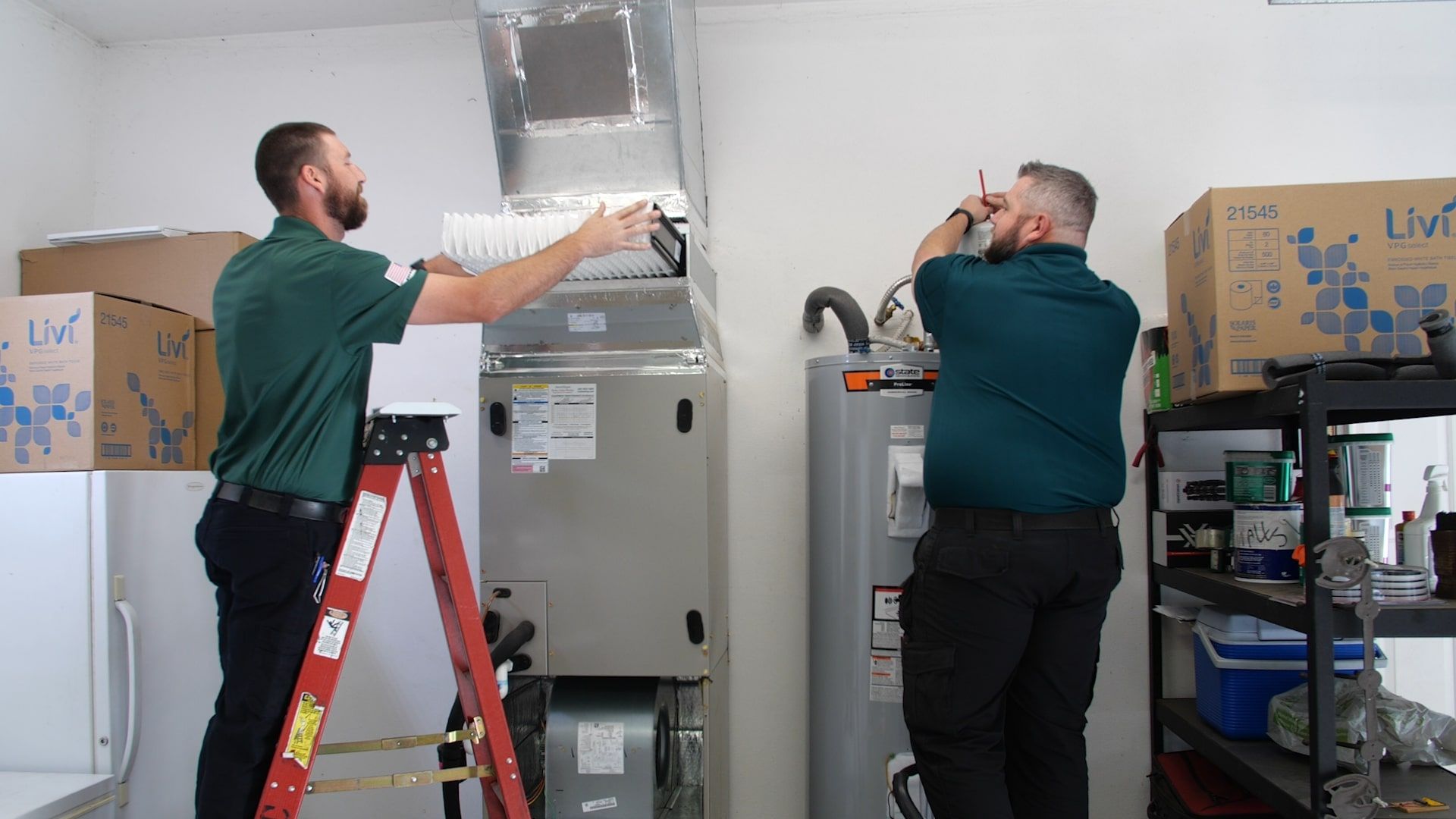 Two HVAC technicians servicing HVAC equipment; one technician is working on the air handler, which houses the air filter and blower, while the other is working on a water heater.